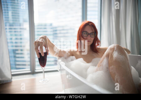 Eine rothaarige Frau im Schaumbad liegen und ein Glas Rotwein halten - Ein Blick auf die modernen Gebäude aus dem Panoramafenster Stockfoto