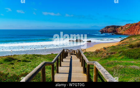 Hölzerne Treppe mit Geländer führt der Weg durch die grüne Gras hinunter zum Strand, Wellen, Himmel, Wolken, viele Menschen auf Surfbrettern, orangefarbene Klippen Stockfoto