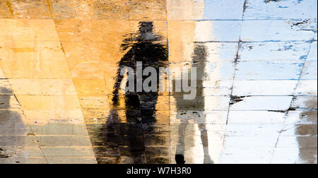 Unscharfe Reflexion Schatten Silhouetten von ein Mann und ein Junge auf einer nassen Straße an einem sonnigen Sommertag in der Altstadt Stein Pflaster Stockfoto