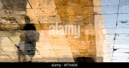 Unscharfe Reflexion Schatten Silhouetten von einer Frau und einem Kind zu Fuß auf einer nassen Straße an einem sonnigen Sommertag auf Altstadt Stein Pflaster Stockfoto