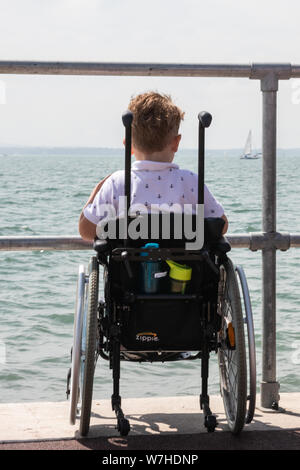 Ein kleiner Junge in einem Rollstuhl am Meer an der Blick auf das Meer suchen Stockfoto