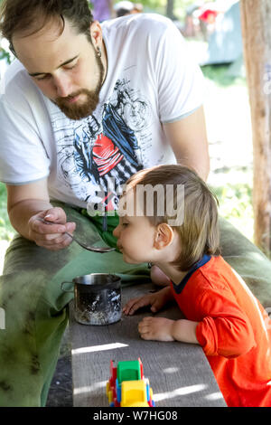 Charkow, Ukraine - Juli 09, 2019: Ein junger Mann zieht ein kleiner Junge mit einem Löffel aus der Tasse, Vater und Sohn im Freien Stockfoto