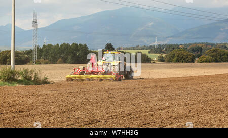 Gelbe Schlepper zieht Rot Aussaat Mechanismus über trockenes Feld, Bäume und Berge im Hintergrund Stockfoto