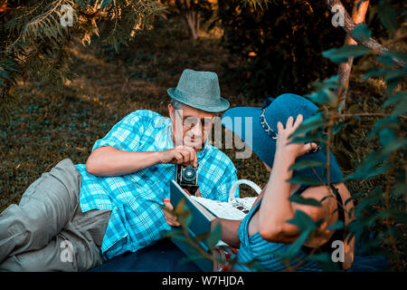Gerne älteres Paar auf dem Picknick mit retro Kamera am Sommer, Park. Ältere Menschen Fotografieren seiner Frau mit einem Buch in den Park Stockfoto