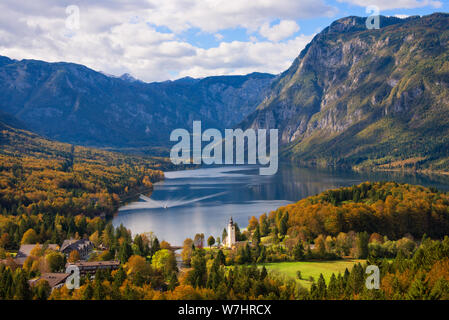 Eine Fähre in Richtung der Stadt Ribčev Laz am See Bohinj und Bäume im Herbst Farben und Julischen Alpen Berge im Herbst Landschaften in Slowenien. Stockfoto