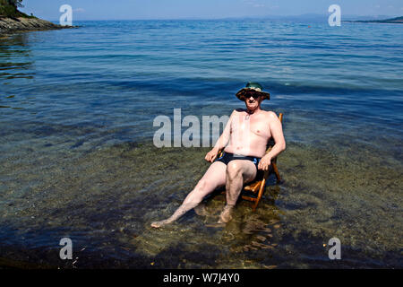 Ein älterer Herr sitzt auf einem Holzstuhl, der sich auf dem Strand in der Ägäis und geniesst die Sonne. Stockfoto