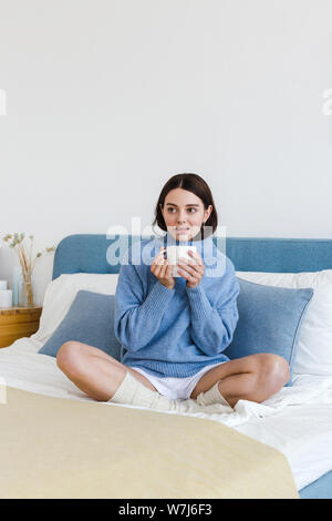 Mädchen in einem blauen Pullover im Innenraum Hygge Stil mit einem Becher in der Hand sitzt auf dem Bett Stockfoto