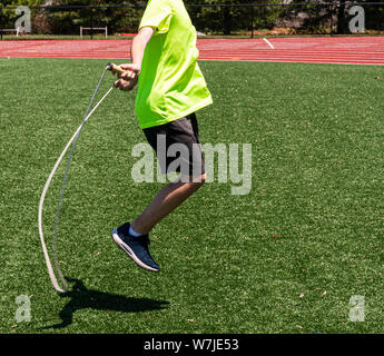 Seite Blick auf eine High School Boy ist Seilspringen auf grünem Rasen Feld tragen ein helles Grün neon Shirt. Stockfoto