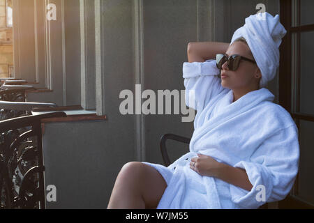 Junge Frau in einem weißen Bademantel, Handtuch und Sonnenbrille in einem Stuhl sitzen auf dem Balkon Terrasse Stockfoto