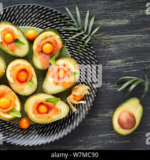 Avocado Boote mit geräuchertem Lachs, Zitrone, Physalis und Mint Blatt. Draufsicht auf dunkel strukturierten Hintergrund, Quadrat Zusammensetzung Stockfoto