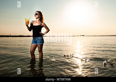 Frau im Meer mit einem Glas Saft mit plastikstrohhalm um viele Kunststoff Flasche schwimmen auf dem Wasser. Umweltschutz Konzept. Stockfoto