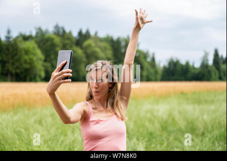 Junge Frau selfies machen die Natur genießen, während sie ihren Arm hebt und macht ein lustiges Gesicht. Mit Fokus auf das Mobiltelefon. Stockfoto