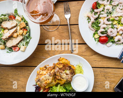 Pikanter salat Garnelen mit Tomaten, Caesar Salat mit Huhn, Salat, Lasagne in weiße Platte mit einem Krug Bier auf Holz- Hintergrund, Abendessen Konzept Stockfoto