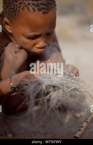 Ein Zu 'hoasi Bushman Kind bläst auf feine Glut in einem Büschel Gras und Pflanzenfasern, um ein Feuer zu machen. Kalahari, Botswana. April 2012. Stockfoto