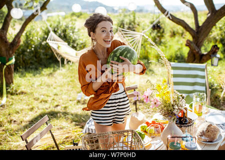 Junge Frau, dekorieren und servieren Tisch, Wassermelone während der Vorbereitung für die festlichen Mittagessen im wunderschönen Garten bei schönem Wetter Stockfoto