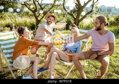 Junge und fröhliche Freunde entspannt auf den Sonnenliegen, klirren Gläser in die wunderschön gestalteten Hinterhof oder im Garten während einer feierlichen Sitzung Stockfoto