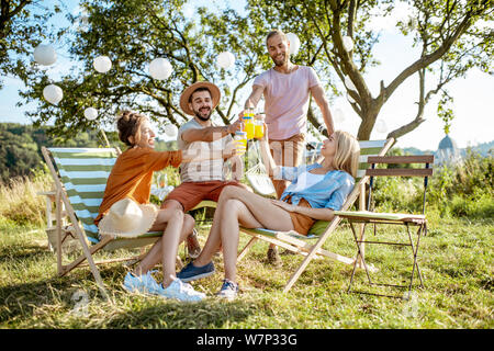 Junge und fröhliche Freunde entspannt auf den Sonnenliegen, klirren Gläser in die wunderschön gestalteten Hinterhof oder im Garten während einer feierlichen Sitzung Stockfoto