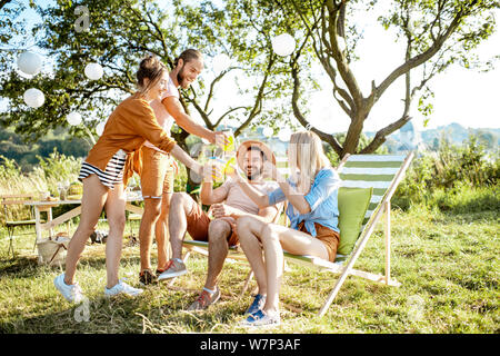 Junge und fröhliche Freunde entspannen mit Getränken, sitzen auf den Sonnenliegen in die wunderschön gestalteten Hinterhof oder im Garten während einer feierlichen Sitzung Stockfoto