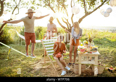 Junge Freunde Spaß haben, zu Fuß auf der Slackline bei einem Picknick in den wunderschön gestalteten Garten auf einen Sonnenuntergang Stockfoto