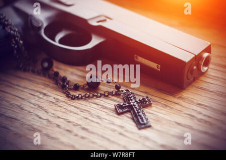 Auf dem alten Holz- Oberfläche liegt eine schwarze Pistole und einem schwarzen Kreuz auf einer Kette mit schwarzen Perlen, die von einem hellen aggressiven Licht beleuchtet. Stockfoto