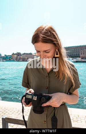 Blonde Schöne junge Mädchen in einem leichten Sommerkleid Grün Khaki Farbe hält eine Kamera und schaut auf digitale Fotos am Meer, an einem sonnigen Tag. Tr Stockfoto