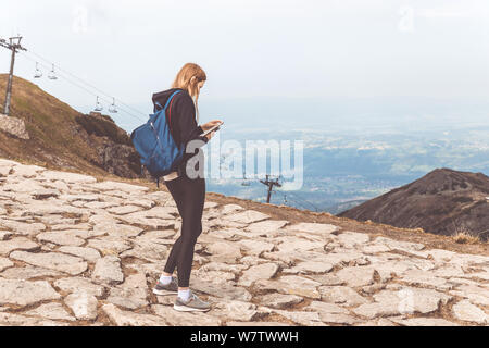 Eine dünne junge Mädchen in einem schwarzen Pullover und Leggings steht in den Bergen und verwendet das Telefon Stockfoto