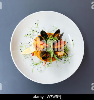 Spanische Paella mit Muscheln und Garnelen mit Zitrone und Rucola. Auf einer weißen Platte mit Zitrone auf einem grauen Tisch serviert. Blick von oben. Stockfoto