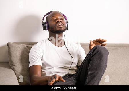 Afrikanische bärtiger Mann sitzen auf der Couch im Wohnzimmer, mit Kopfhörer auf dem Kopf und seine Lieblingsmusik hören Stockfoto