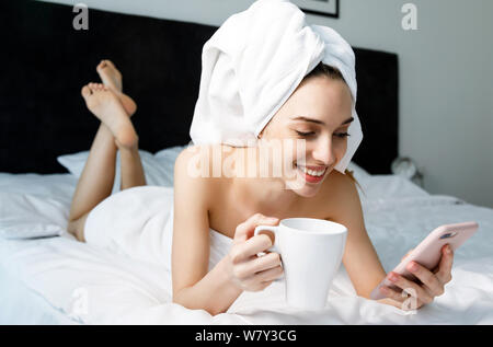 Schöne Frau mit Handtuch auf dem Kopf schreiben Nachricht und Trinken moning Kaffee im Schlafzimmer zu Hause Stockfoto