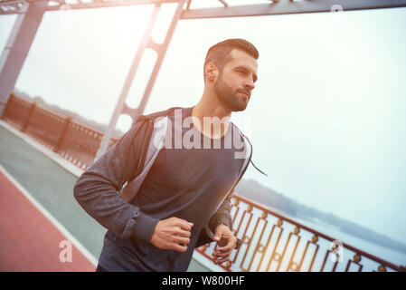 Stark und gesund. Seitliche Sicht auf junge, gesunde, bärtigen Mann im Sport Kleidung Jogging auf der Brücke. Sport Konzept. Gesunder Lebensstil Stockfoto