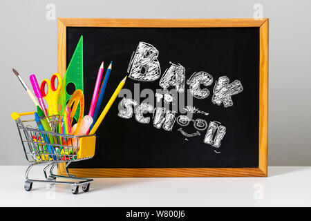 Verschiedenen bunten Schulmaterial und Einkaufswagen auf dem Hintergrund der School board mit den Worten Zurück zur Schule Stockfoto