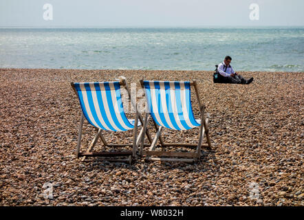 Zwei blau-weiß gestreifte Liegestühle am Kieselstrand mit Person im Hintergrund, Brighton, East Sussex UK Stockfoto