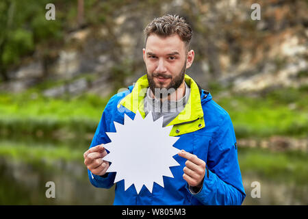 Eine freche junge Mann ist eine Karte leer für Ihre Werbung in seinen Händen halten, er steht auf einer unscharfen natürliche Landschaft. Stockfoto