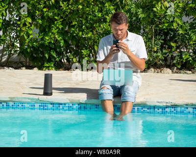 Ein junger Mann in Jeans und Shirt ist die Arbeit mit einem Laptop auf das Internet und auf ein Handy in der Nähe des Pools. Stockfoto