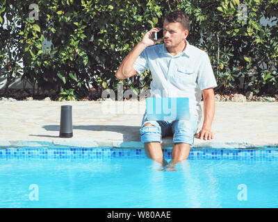 Ein junger Mann in Jeans und Shirt ist die Arbeit mit einem Laptop auf das Internet und auf ein Handy in der Nähe des Pools. Stockfoto