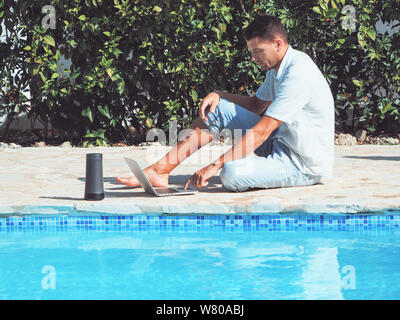 Ein junger Mann in Jeans und Shirt ist die Arbeit mit einem Laptop im Internet in der Nähe der Pool. die Füße im Wasser. Stockfoto