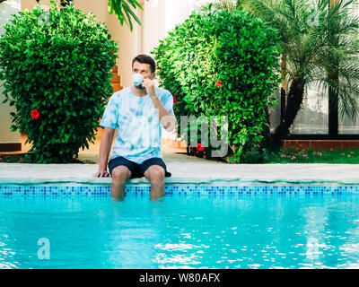 Portrait von reifem Alter Mann, der ein T-Shirt und Shorts in der Nähe ein Schwimmbad trinken Kaffee sitzen. Füße im Pool im Wasser. Stockfoto