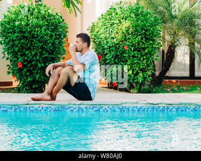 Portrait von reifem Alter Mann, der ein T-Shirt und Shorts in der Nähe ein Schwimmbad trinken Kaffee sitzen. Stockfoto