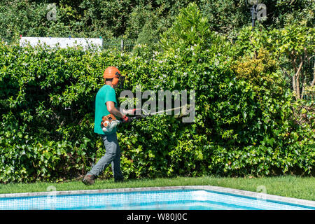 Ein Mann mit einem freischneider ausgestattet arbeitet auf Vorgarten mit Swimmingpool. Garten Wartungskonzept Stockfoto