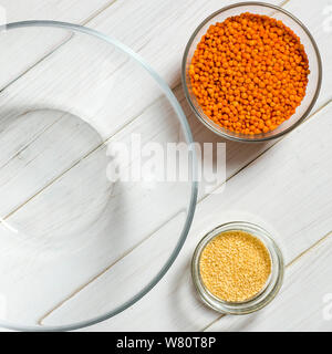 Glasplatten mit roten Linsen, Sesamsamen und eine große Glasplatte stehen auf einem weißen Tisch Stockfoto