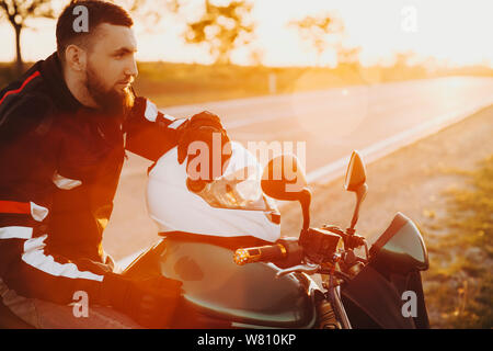 Seitenansicht Porträt einer jungen Bartgeier kaukasischen Mann sitzt auf seinem Fahrrad mit einer Hand auf seinen Helm und nachdenklich auf der Suche gegen Sonnenuntergang während t Stockfoto