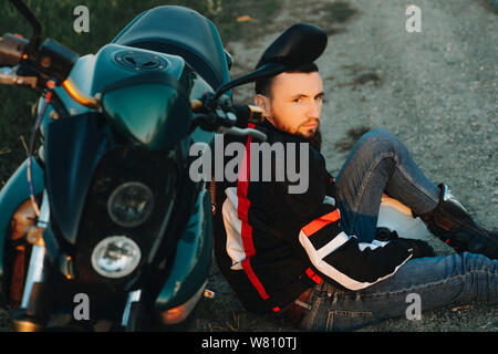 Portrait eines bärtigen zuversichtlich Mann in den Boden lehnte sich auf seinem Motorrad Weg ernsthaft beim Erforschen der neuen Plätze in seinem Weg sitzen. Stockfoto