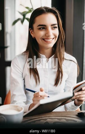 Portrait einer jungen attraktiven kaukasischen Geschäftsfrau, sitzen in einem Café am Mittag und schreiben in Ihrem Notebook weg schauen lächelnd. Stockfoto