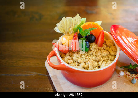 Apple Crumble in kleinen Keramik orange Schimmel Topf und Spitze mit Obst, Erdbeere, Orange und Black, und die Creme auf die Seite, auf Holz- Fach und Tisch. Stockfoto