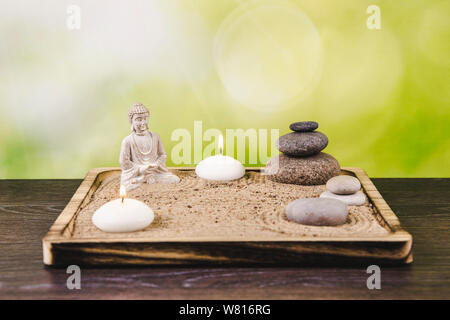 Miniatur Schreibtisch zen Sandbox mit Buddha Figur sitzen im Lotussitz, gestapelte zen Meer Steine, Wellness Kerzen brennen gegen grüne Bokeh studio Hintergrund Stockfoto