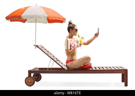 Volle Länge Profil Schoß einer Frau im Bikini ein selfie Foto und trinken Cocktail auf einem Liegestuhl unter dem Sonnenschirm auf weißem Hintergrund Stockfoto