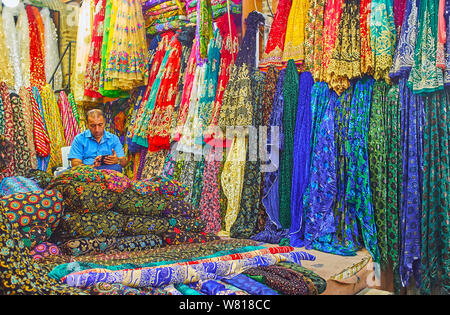 SHIRAZ, IRAN - Oktober 14, 2017: Der Kaufmann in Textil- stand von vakil Basar unter bunten Stoffen, eingerichtet mit Mustern, Stickereien und Lurex Stockfoto