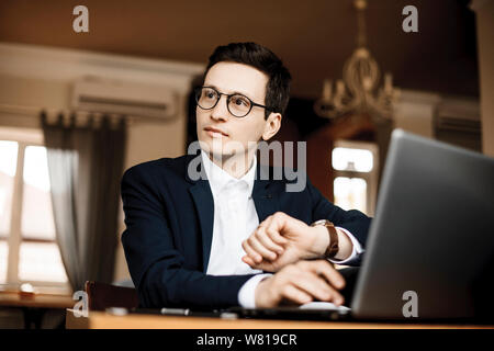 Porträt einer hübscher junger Freelancer Brille weg, während auf seine Uhr im Coffeeshop an seinem Laptop arbeiten sitzen auf der Suche suchen. Stockfoto