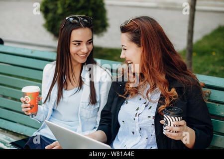 Zwei reizende junge Freundinnen trinken Kaffee beim Sitzen auf einer Bank mit einem Laptop und einem Tablet suchen einander lachend mit einer Tasse heißen Kaffee. Stockfoto