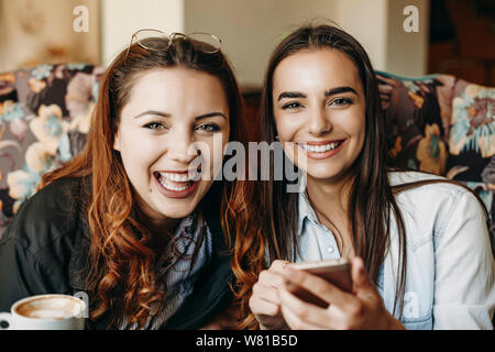 Portrait von zwei schönen weiblichen Kamera schaut lachend während man ein Smartphone in der Hand halten, während in einem Coffee Shop in Ihren vaca Sitzen Stockfoto